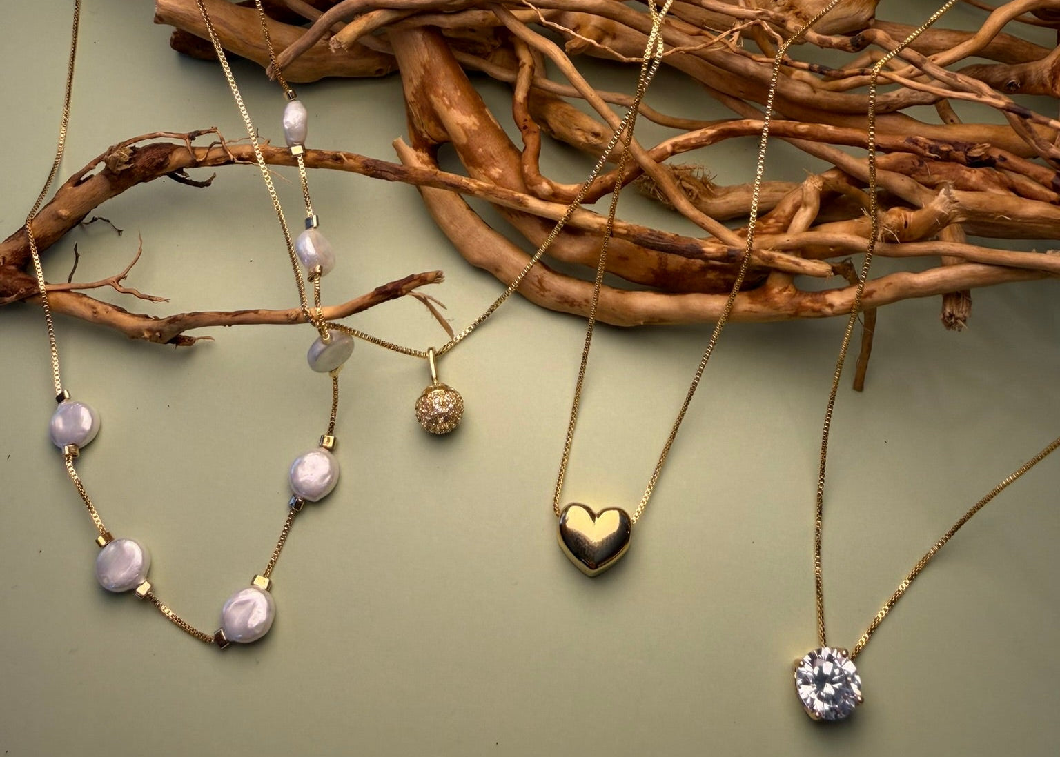 Several delicate gold-tone necklaces with various pendants are draped over a rustic arrangement of natural driftwood branches against a muted sage green background. The necklaces feature different designs including a heart-shaped pendant, a cluster of light purple pearls, and a clear faceted gemstone.